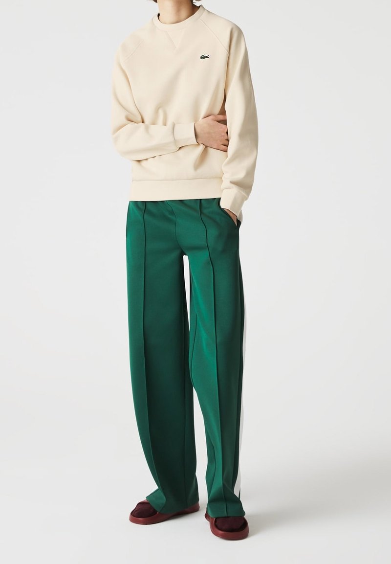 Sudadera beige con puños acanalados, combinada con pantalones de pierna ancha verdes que tienen pliegues laterales y acentos blancos. Unos zapatos marrones de slip-on completan el look.