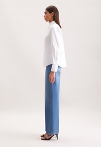 Camicia bianca con bottoni e maniche lunghe abbinata a jeans blu a vita alta e a gamba dritta, indossata con tacchi a stiletto neri, mostrata di lato.