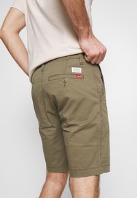 Pantaloni corti verde oliva realizzati in tessuto misto cotone, con una tasca posteriore con bottone e un'etichetta con marchio cucita sulla vita.