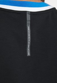 Närbild av en svart t-shirt med en ryggkrage med blå och vit trim och en vertikal grå remsa med texten "LÅT INTE BRA STÅ I VÄGEN FÖR STORT."