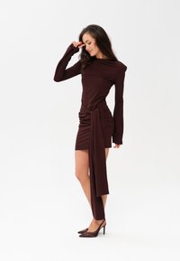 Ciemnobrązowa sukienka z długim rękawem, z marszczeniem w pasie, asymetrycznym krojem i opadającym akcentem z tkaniny. Połączona z czółenkami na szpilce.