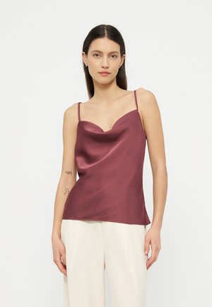 Jauna moteris su ilgais tamsiais plaukais dėvi rankovių neturintį bordo satino viršų ir baltas platesnes kelnes, stovėdama priešais šviesų vienodą foną.