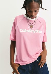 Person iført en lyserød "Calm Rhythm" T-shirt, lagdelte sølvkæder med vedhæng af et hjerte, mørke jeans og flere ringe, som ser nedad.