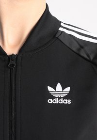 Černá sportovní bunda s vysokým límcem, obsahující bílé logo Adidas a tři bílé pruhy podél ramen. Hladký, přiléhavý materiál.