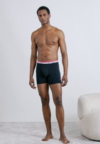 Μαύρα μπόξερ σορτς από ελαστικό ύφασμα. Διαθέτει ροζ και λευκή ζώνη μέσης με το λογότυπο της Nike. Το μοντέλο στέκεται σε ανοιχτόχρωμη μοκετά.