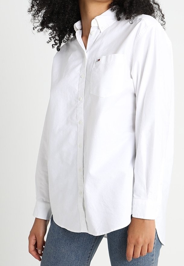 Femme portant une chemise blanche boutonnée avec une poche avant et un jean bleu, debout devant un fond uni.