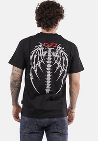 Uomo con capelli ricci e tatuaggi che indossa una maglietta nera con un design di spina dorsale e ali di drago bianco, con fiamme rosse sul retro.