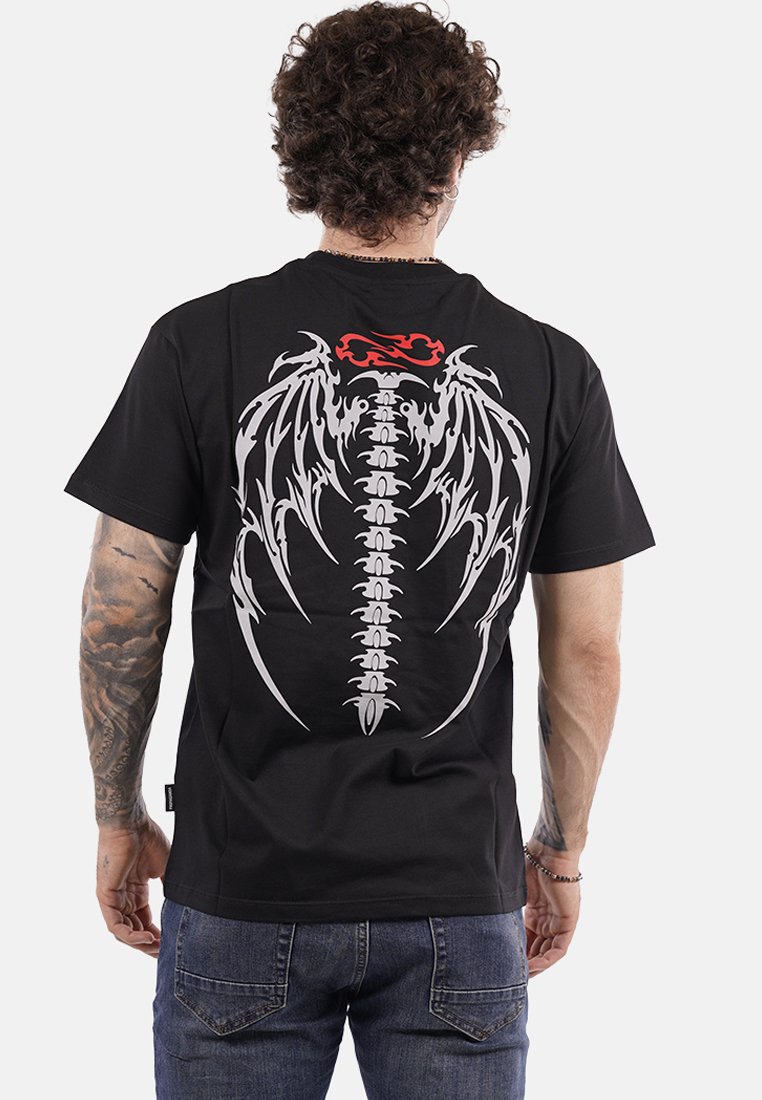 Uomo con capelli ricci e tatuaggi che indossa una maglietta nera con un design di spina dorsale e ali di drago bianco, con fiamme rosse sul retro.