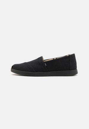 Sort slip-on lærredsko med flad sål, lav profil og lille sidetag, vist fra den indvendige side på en hvid baggrund.