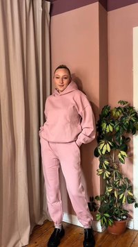 Młoda osoba stoi z rękami w kieszeniach, ubrana w jasnoróżową bluzę z kapturem i dopasowane spodnie obok zielonej, liściastej rośliny na tle różowej ściany.