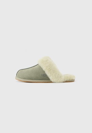 Pantofole in suede verde chiaro con foderatura e bordo in morbido color crema, marchiate con il logo UGG sul lato, dotate di suola piatta.