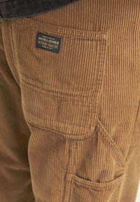 Brown corduroy pants featuring a prominent stitched texture, side pocket, and a blue brand label with white text.