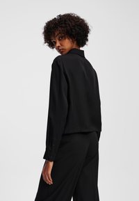 Camicia nera a maniche lunghe con colletto, tessuto morbido, polsini con bottoni e design cropped. Il retro è semplice, evidenziando uno stile minimalista.