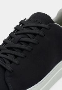 Gros plan d'une basket noire avec des lacets noirs et une semelle blanche, montrant la languette avec un marquage en relief subtil.