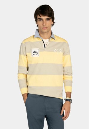 Joven con una camiseta de rugby a rayas amarillas y beige con cuello azul, pantalones azules y un reloj en la muñeca, de pie con una mano en el bolsillo.