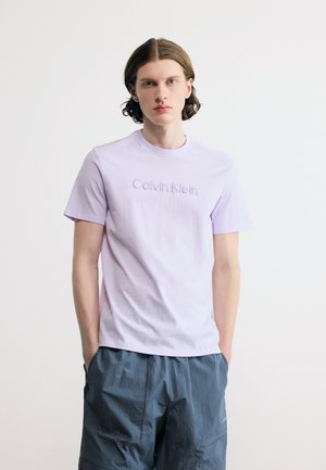 Bărbat tânăr cu păr până la umeri poartă un tricou Calvin Klein de culoare lavandă deschisă și pantaloni scurți albastru închis, stând în picioare pe un fundal simplu alb.