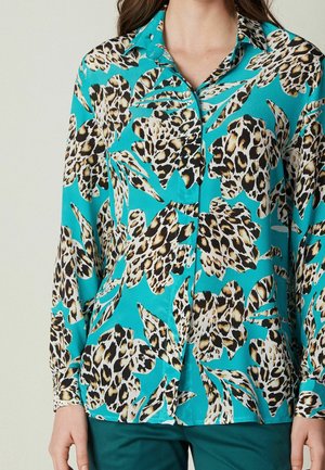Turquoise button-up shirt with long sleeves and large leopard print leaf patterns worn with dark green pants.