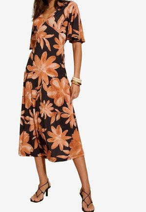 Femme portant une robe midi noire avec un grand motif floral orange, des manches courtes et des sandales noires à talons hauts avec brides.