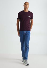 Bordeaux kortærmet t-shirt med "Lee" logo på venstre bryst, parret med blå jeans og grå-grønne sneakers. Enkel, tætsiddende design.