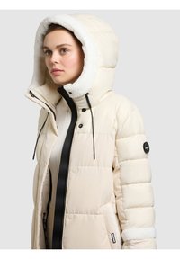 Vadderad jacka i krämfärg med luva fodrad med vit fleece, svart dragkedja och sidofickor med detaljer i pälsliknande textur.