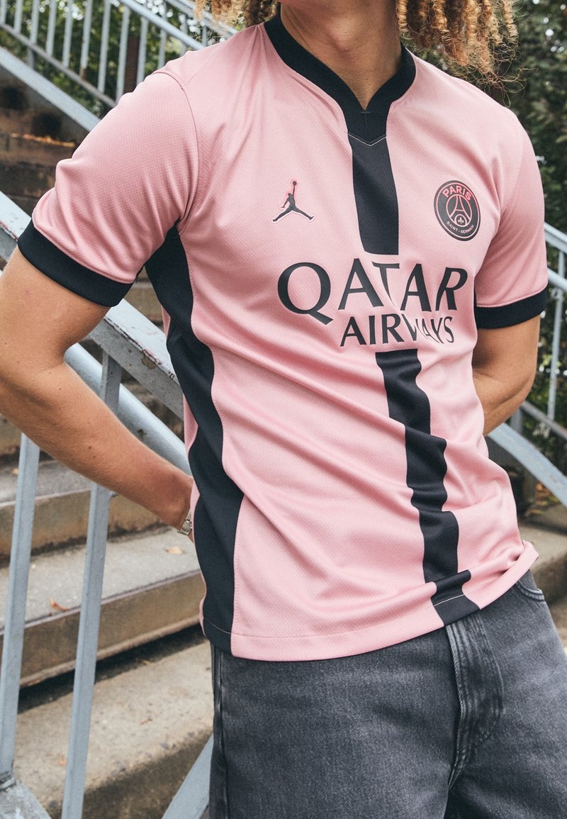 Osoba oblečená v růžovo-černém fotbalovém dresu Paris Saint-Germain s logem "Qatar Airways", stojící vedle venkovního kovového schodiště.