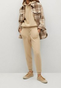 Beige Kapuzenpullover mit Kordelzug, passende tapered Jogginghosen und ein braunes karriertes Flanellhemd. Tanfarbene Sneakers runden das Outfit ab.