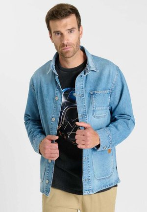 Uomo che indossa una giacca in denim azzurro chiaro sopra una maglietta nera con stampa grafica, pantaloni beige, in piedi davanti a uno sfondo bianco semplice.