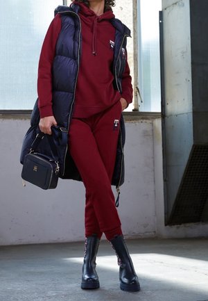 Sudadera con capucha y pantalones deportivos color granate combinados con un chaleco acolchado azul marino. Botines negros con detalles decorativos. Pequeño bolso negro con herrajes metálicos.