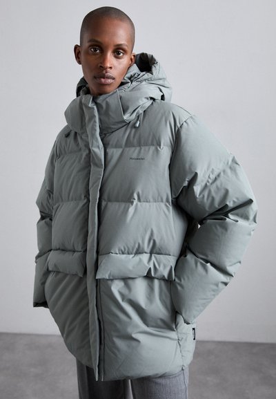 Padded parka in soft, light gray with a high collar, adjustable hood, and front pockets. Features logo on the chest and a textured finish.