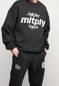 Sudadera negra de gran tamaño con texto gráfico blanco y pantalones deportivos negros a juego que presentan detalles de logo y bolsillos. Hecho de tejido suave.