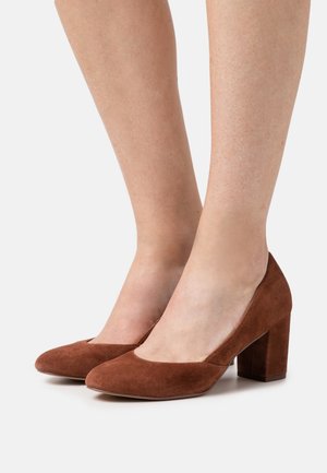 Escarpins en daim marron à talons hauts avec un bout arrondi et un talon carré. Les chaussures présentent une texture lisse et une couleur unie sans motifs.