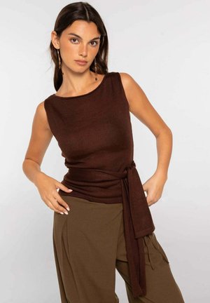 Mujer con cabello largo y oscuro, que lleva una blusa marrón sin mangas con la cintura anudada y pantalones marrones de cintura alta, de pie frente a un fondo liso.