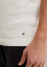 Fehér textúrájú kötött pulóver bordás aljjal, oldalon egy kis fényes logóval, fekete nadrággal párosítva.