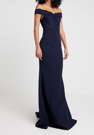 Vrouw draagt een marineblauwe off-shoulder jurk met kanten lijfje en lange aansluitende rok, staand tegen een effen achtergrond.