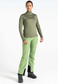 Langärmliges grünes Oberteil mit halbem Reißverschluss, kombiniert mit hellgrünen Hosen, die schwarze Akzente an den Säumen aufweisen, und schwarzen Wanderschuhen.