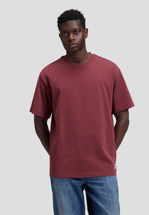 Jeune homme aux cheveux courts et bouclés, portant un t-shirt uni bordeaux à manches courtes et un jean bleu, se tenant devant un fond gris.