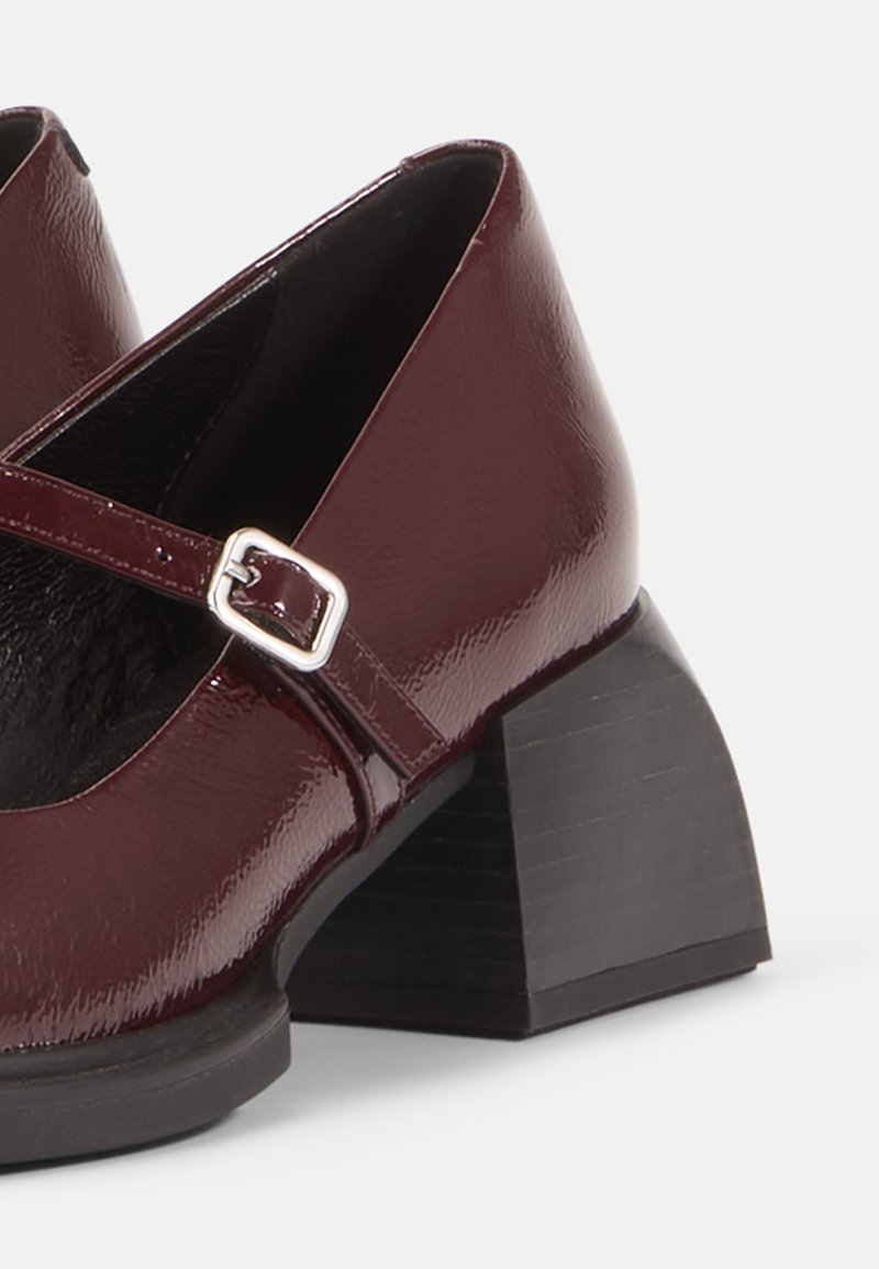 Scarpe in pelle verniciata bordeaux con cinturino incrociato e fibbia argentata; tacco quadrato nero con superficie testurizzata.
