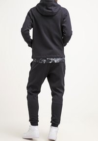 Sudadera negra con capucha y joggers a juego, con un tejido texturizado, corte relajado y zapatillas deportivas blancas. Vista posterior.