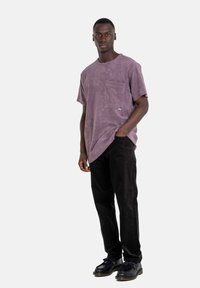 Lila Baumwoll-T-Shirt mit einer Brusttasche, kombiniert mit schwarzen Cordhosen. Das Model trägt schwarze Schuhe mit glänzender Oberfläche.