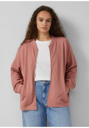 Giacca bomber rosa cipria con zip, in tessuto testurizzato, maniche a coste e tasche frontali inclinati, abbinata a una t-shirt bianca e jeans blu chiaro.