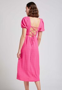 Robe rose mi-longue avec des manches bouffantes, tissu texturé, et un design de dos en croix avec un détail de lien.