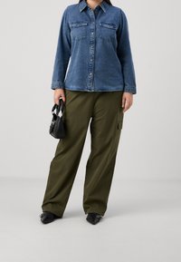 Denim button-up shirt in dark blue with two chest pockets, paired with olive green wide-leg pants and black pointed shoes, holding a small black bag.