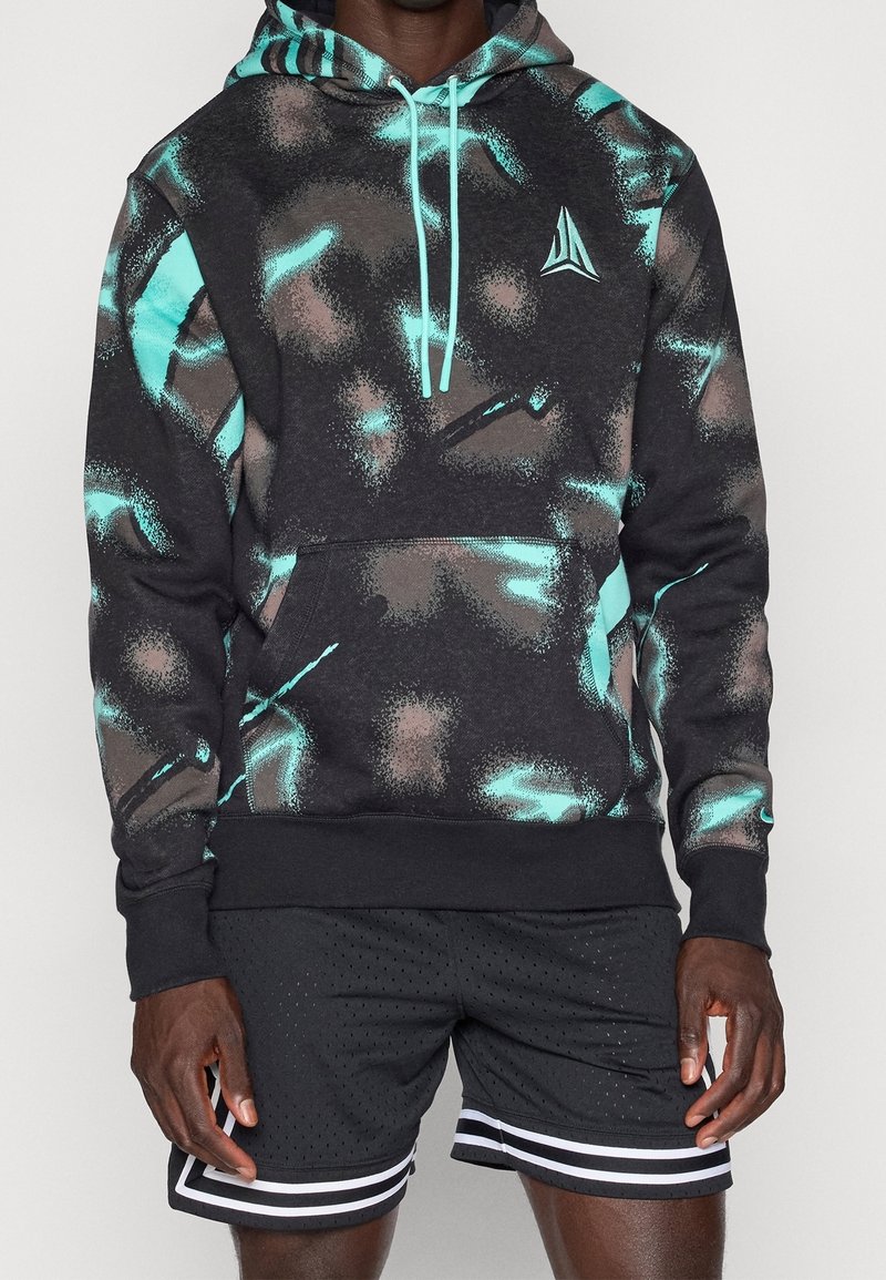 Homme portant un sweat à capuche noir avec des motifs abstraits turquoise et marron et un short de sport noir avec des rayures blanches.