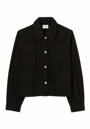 Donna con lunghi capelli scuri che indossa una giacca nera con cintura, pantaloni neri coordinati e scarpe marroni, in piedi davanti a uno sfondo chiaro uniforme.