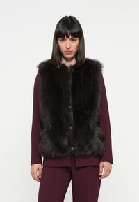 Gilet en fausse fourrure noire avec un devant à boutons, des poches latérales et un col, superposé sur un haut à manches longues bordeaux et un pantalon assorti.