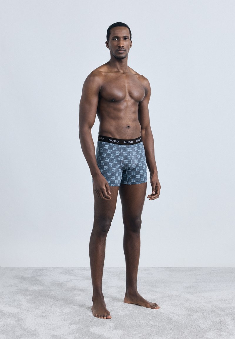 Cuecas boxer masculinas com padrão cinzento apresentando o logótipo "HUGO" repetido. Tecido macio, ajuste justo e cintura elástica preta. Figura em pé, descalço.