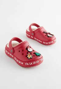 Röda träskor med mjukt innanför, prydda med julinspirerade charms (tomte, gran, pepparkaka) och texten "FA LA LA LA" runt sulan.