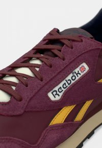 Close-up de um sneaker Reebok cor vinho com textura de camurça, faixas laterais amarelas e atacadores vinho em fundo branco.