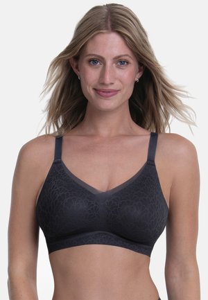 Femme aux cheveux blonds portant un soutien-gorge sans armatures gris foncé à motifs avec des bretelles réglables, souriante sur un fond uni.
