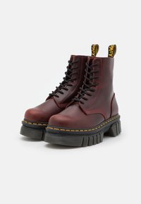 Botas de tobillo de cuero rojo con cordones negros, suela de goma negra gruesa, costuras amarillas y un lazo de marca en la parte superior.